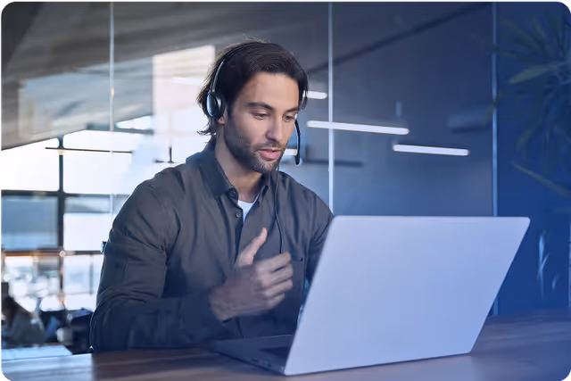 Mann mit Headset spricht vor einem aufgeklappten Laptop und erklärt mit einer Handbewegung etwas